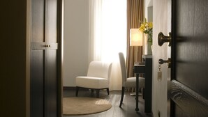A hotel room with a wooden floor, a white chair, a desk with a lamp, and a window with curtains. - Romeo al Babuino (Rome)