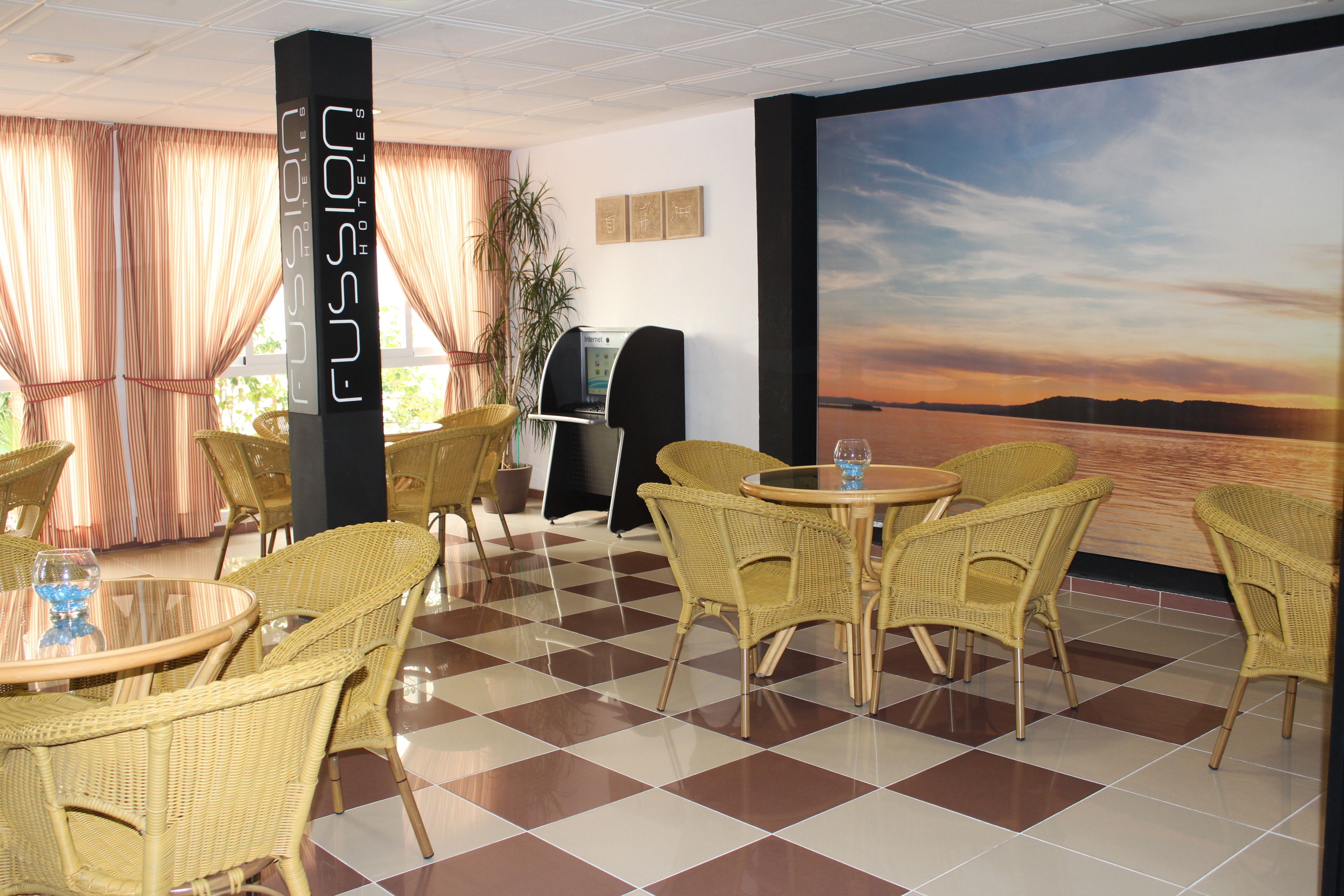 Un vestíbulo de hotel con mobiliario de mimbre, piso a cuadros y un gran mural en la pared que muestra un atardecer sobre el agua.