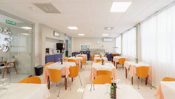 A dining area with tables set for meals, chairs, and a kitchen area in the background.