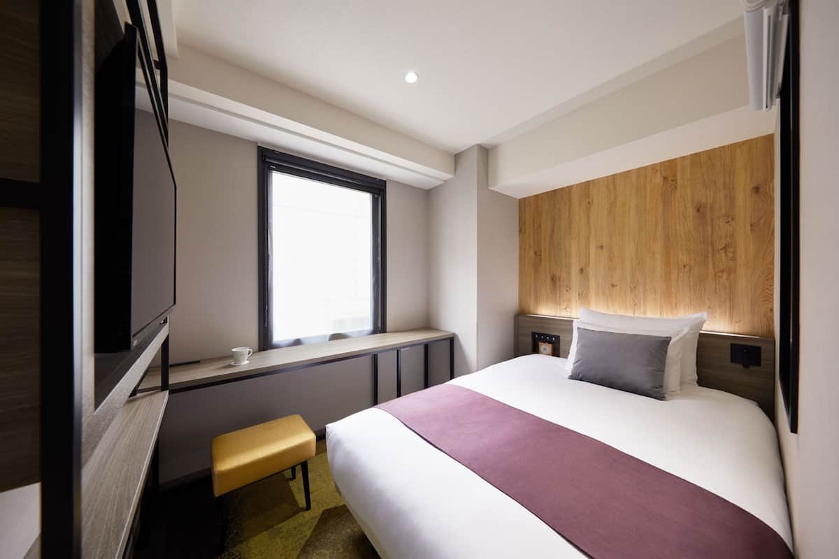 Una habitación de hotel moderna con una cama grande, cabecera de madera, una ventana pequeña, un escritorio con silla y televisor.