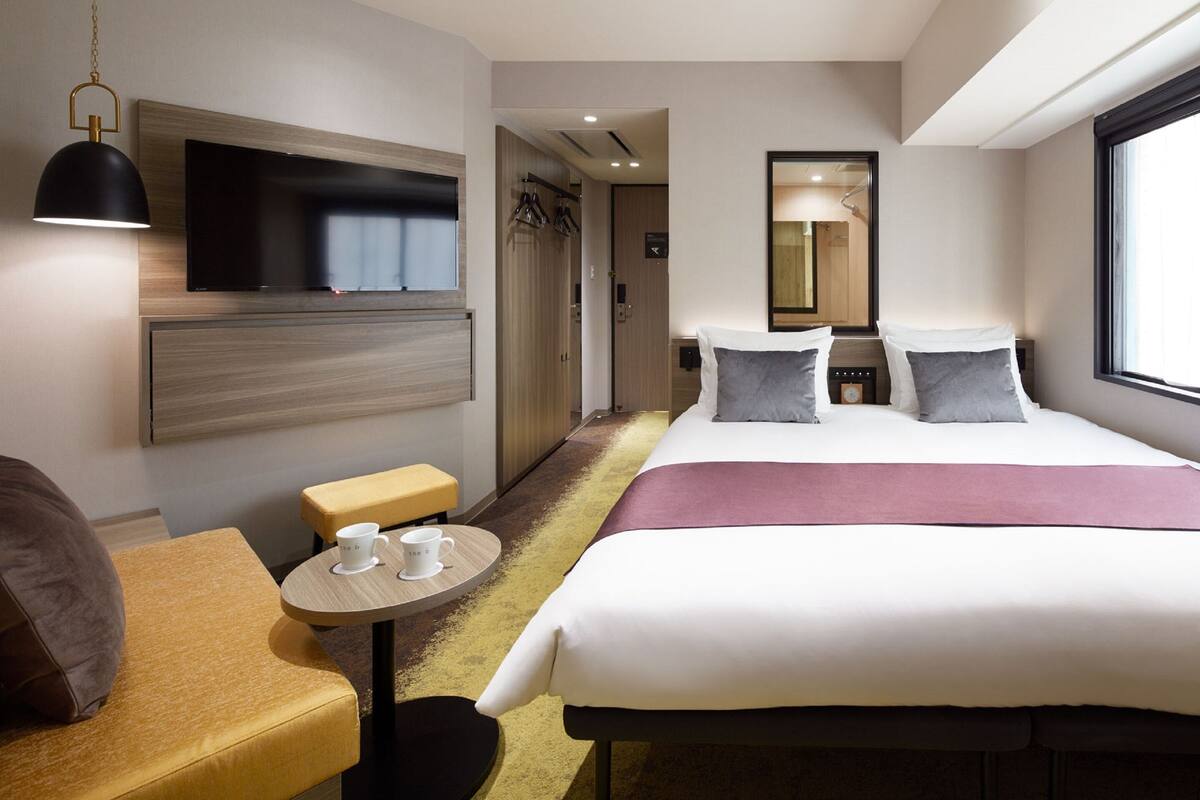 Habitación de hotel con una cama grande, un televisor de pantalla plana montado en la pared, una mesita de noche con una taza, un pequeño rincón de descanso con un banco y una ventana que proporciona luz natural.