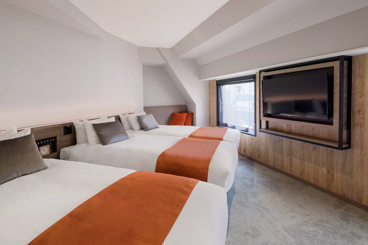 Habitación de hotel con tres camas, un televisor montado en la pared y un ventanal que proporciona luz natural.
