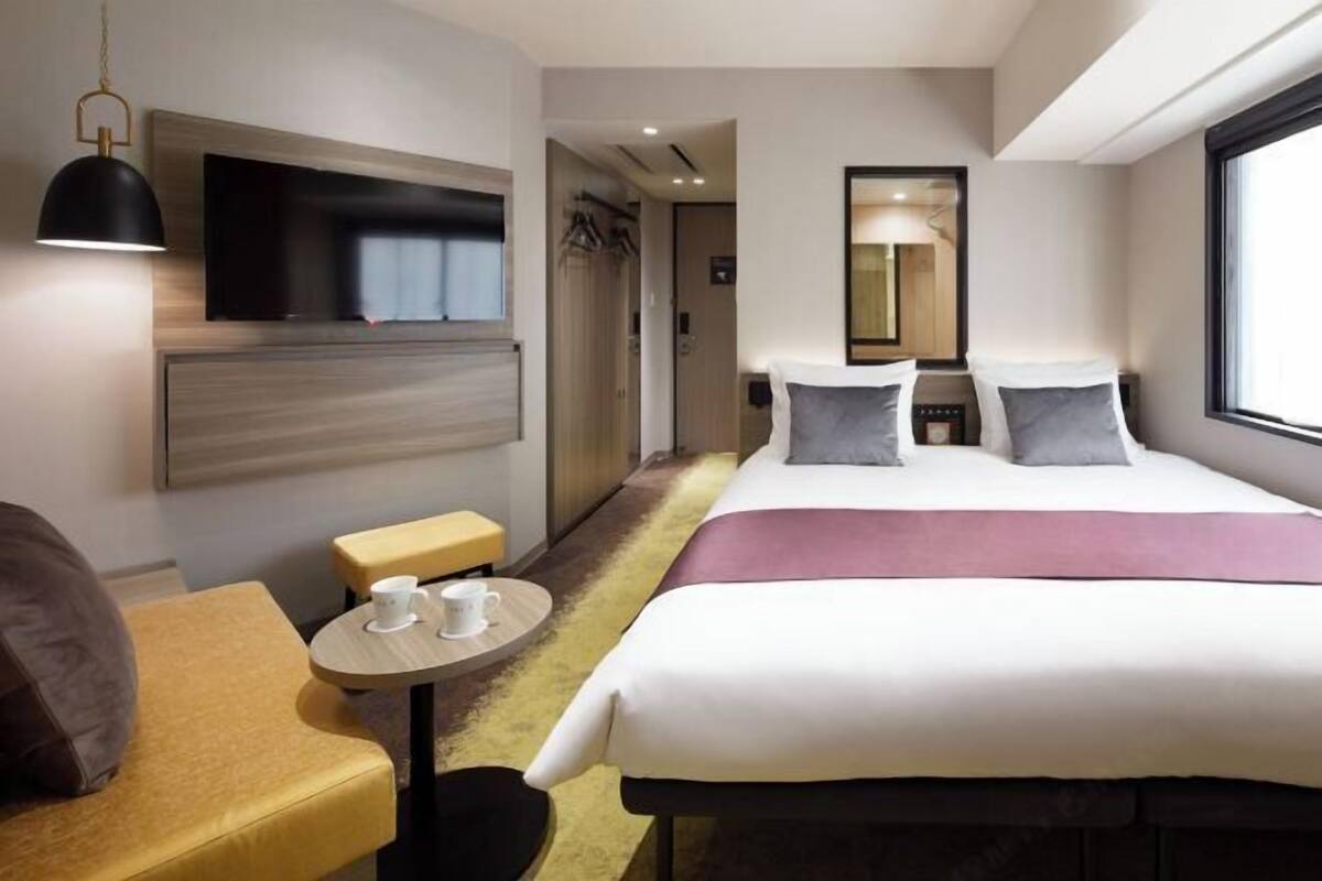 Habitación de hotel con cama, mesita de noche, televisor en la pared, espejo y ventana.