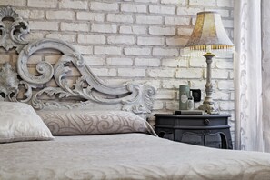 A bedroom with a white brick wall, ornate bed headboard, a bedside table with a lamp, and a nightstand. - Bloom (Venice)