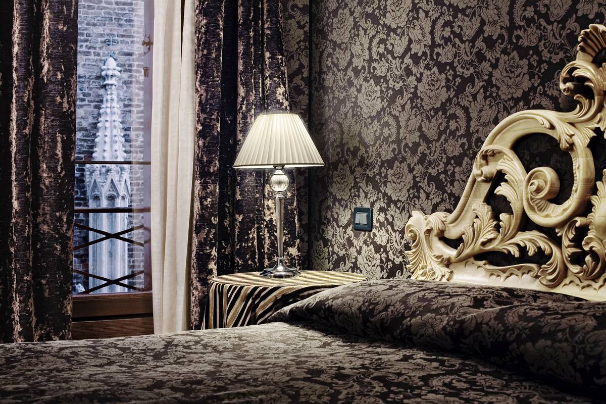 A bedroom with a large ornate bed, a bedside table with a lamp, and a view of a building through a window with curtains.