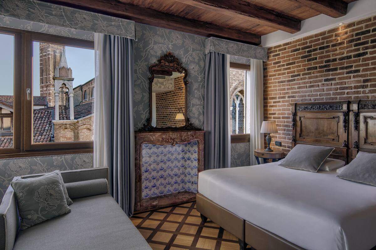A room with a bed, a sofa, a fireplace, and a large window with a view of a brick building.