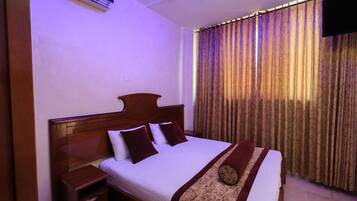 Habitación de hotel con cama de madera, ropa de cama blanca, una mesita de noche, televisor montado en la pared y una ventana con cortinas estampadas.