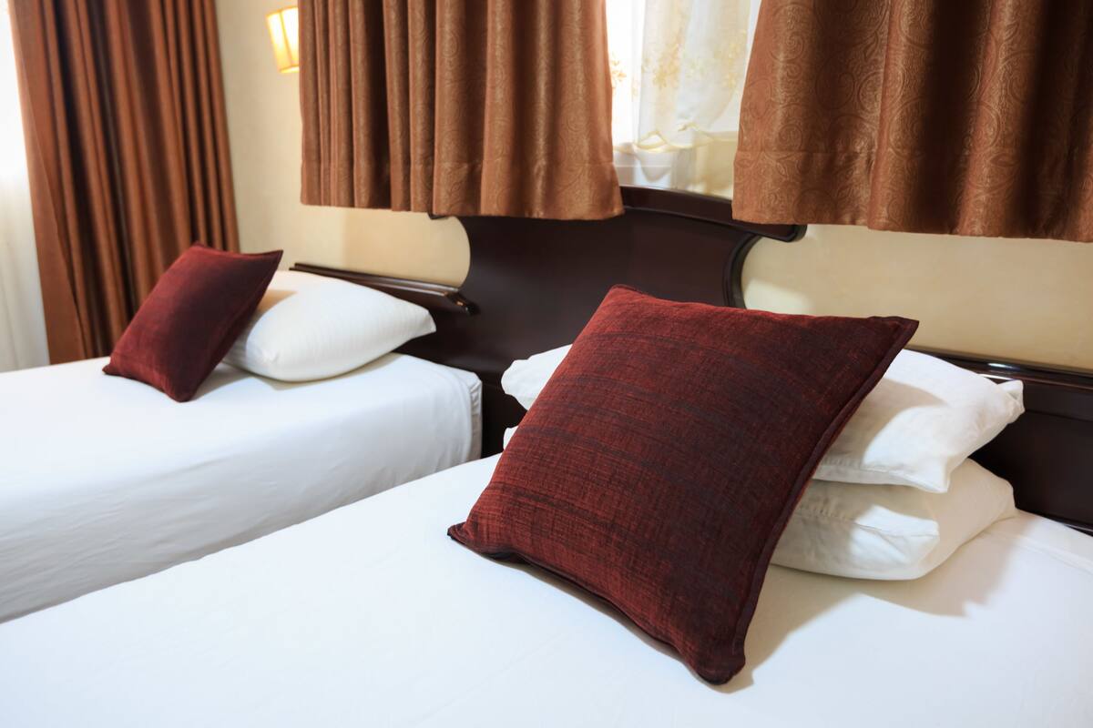 Two single beds with white linens and red pillows, brown curtains, and a wooden headboard.