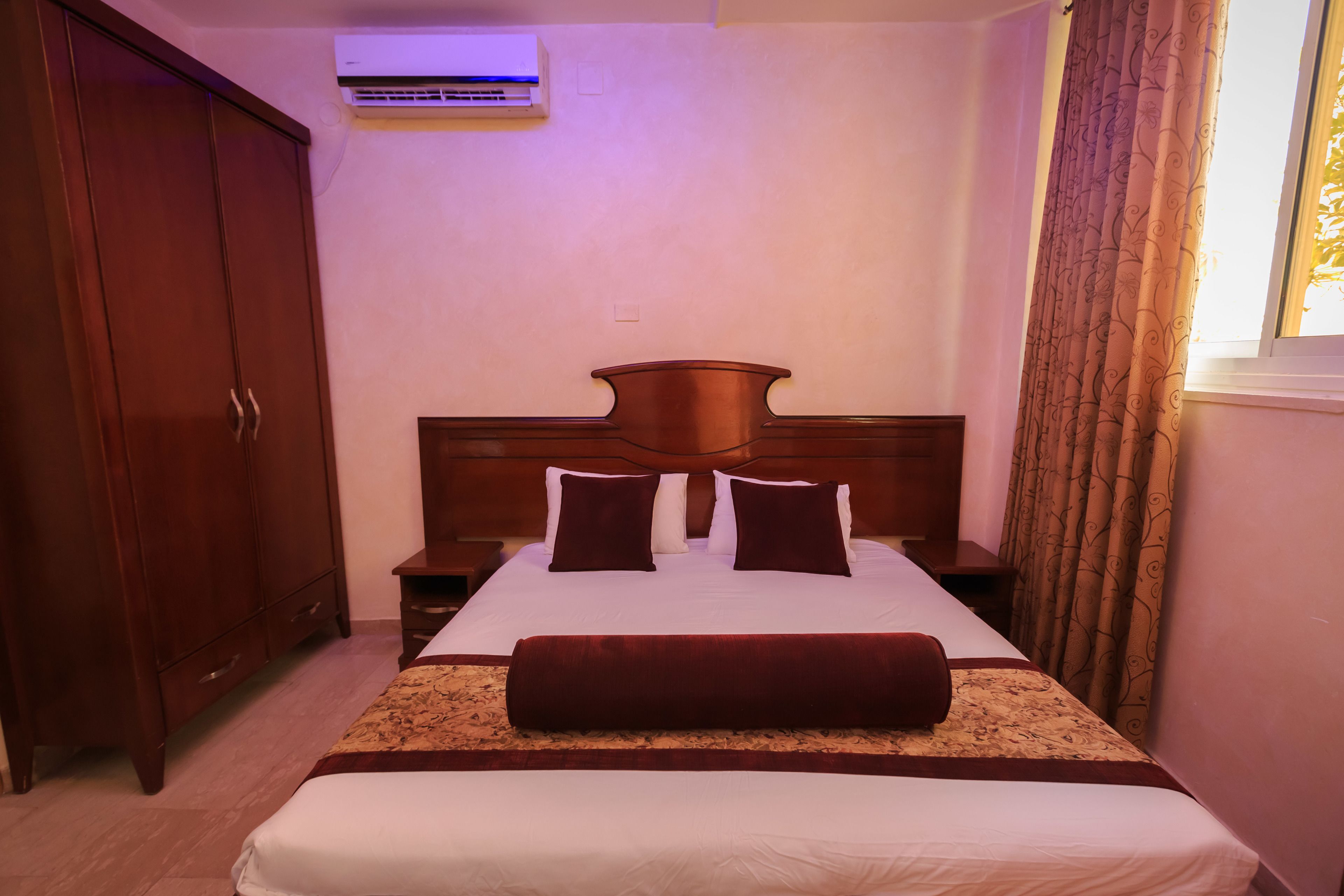 A neatly made bed with a headboard, a wooden wardrobe, and a window with curtains.