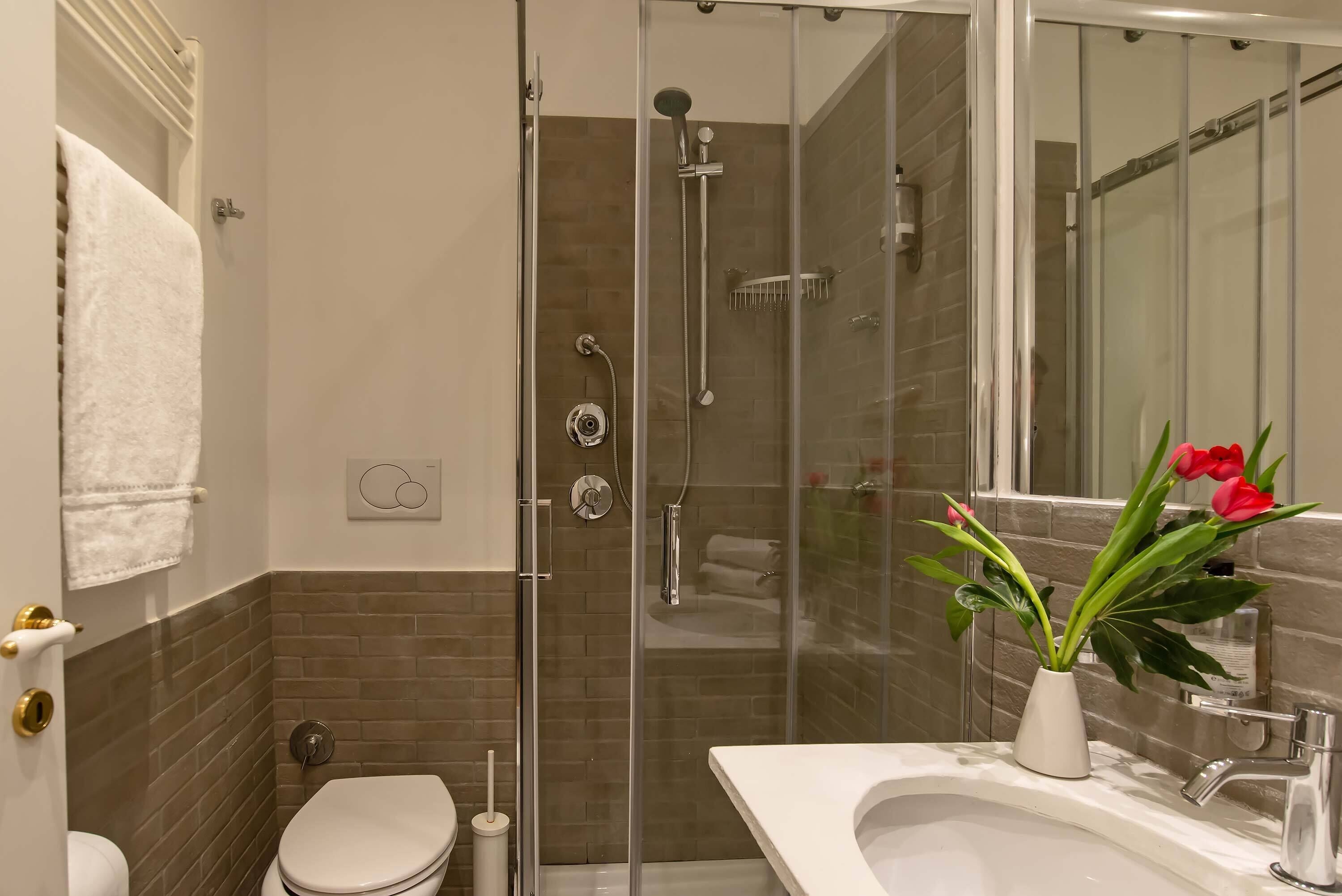 A bathroom with a glass-enclosed shower, a toilet, and a sink with a vase of flowers.