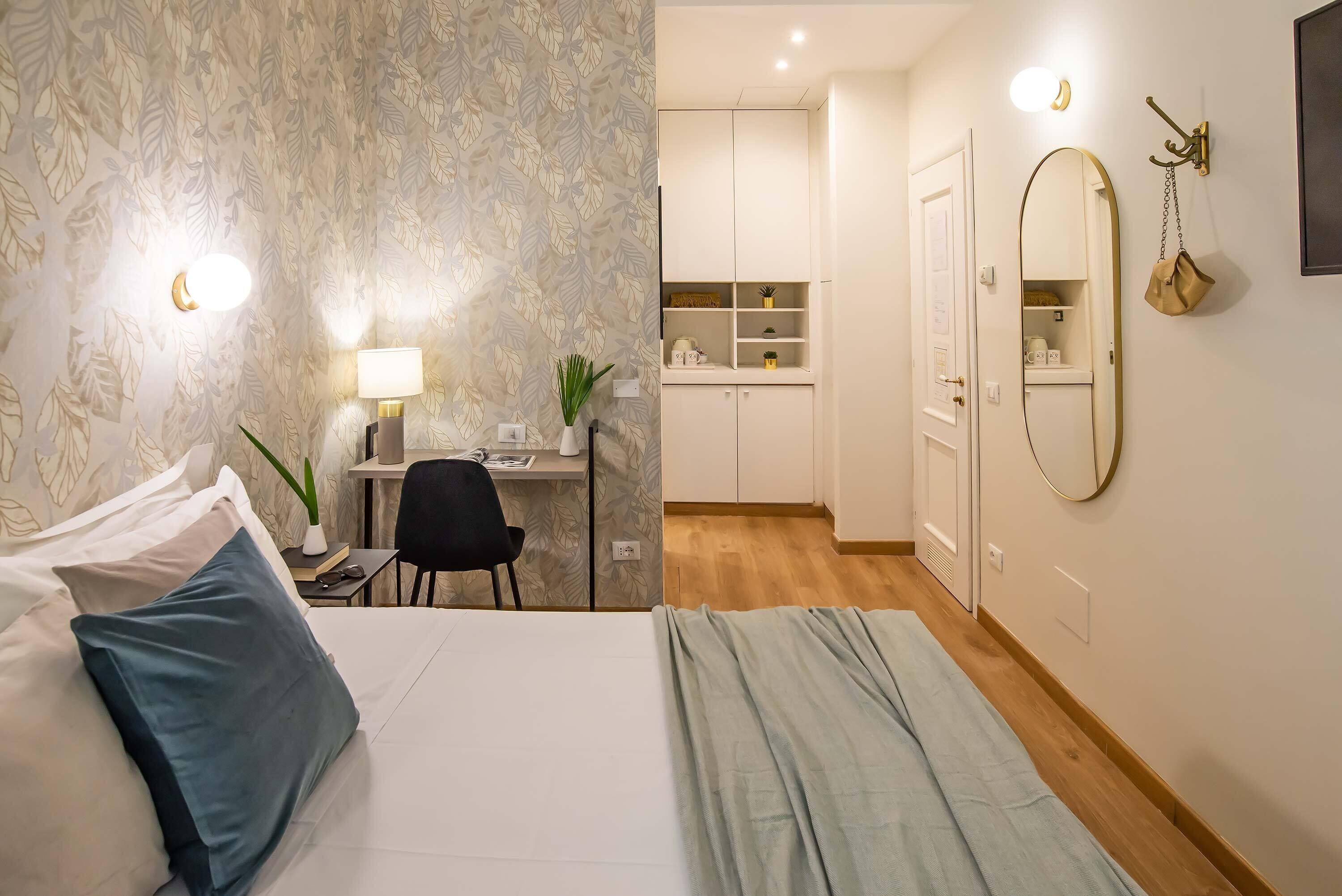 A hotel room with a bed, a desk with a chair, a mirror, and a wardrobe.
