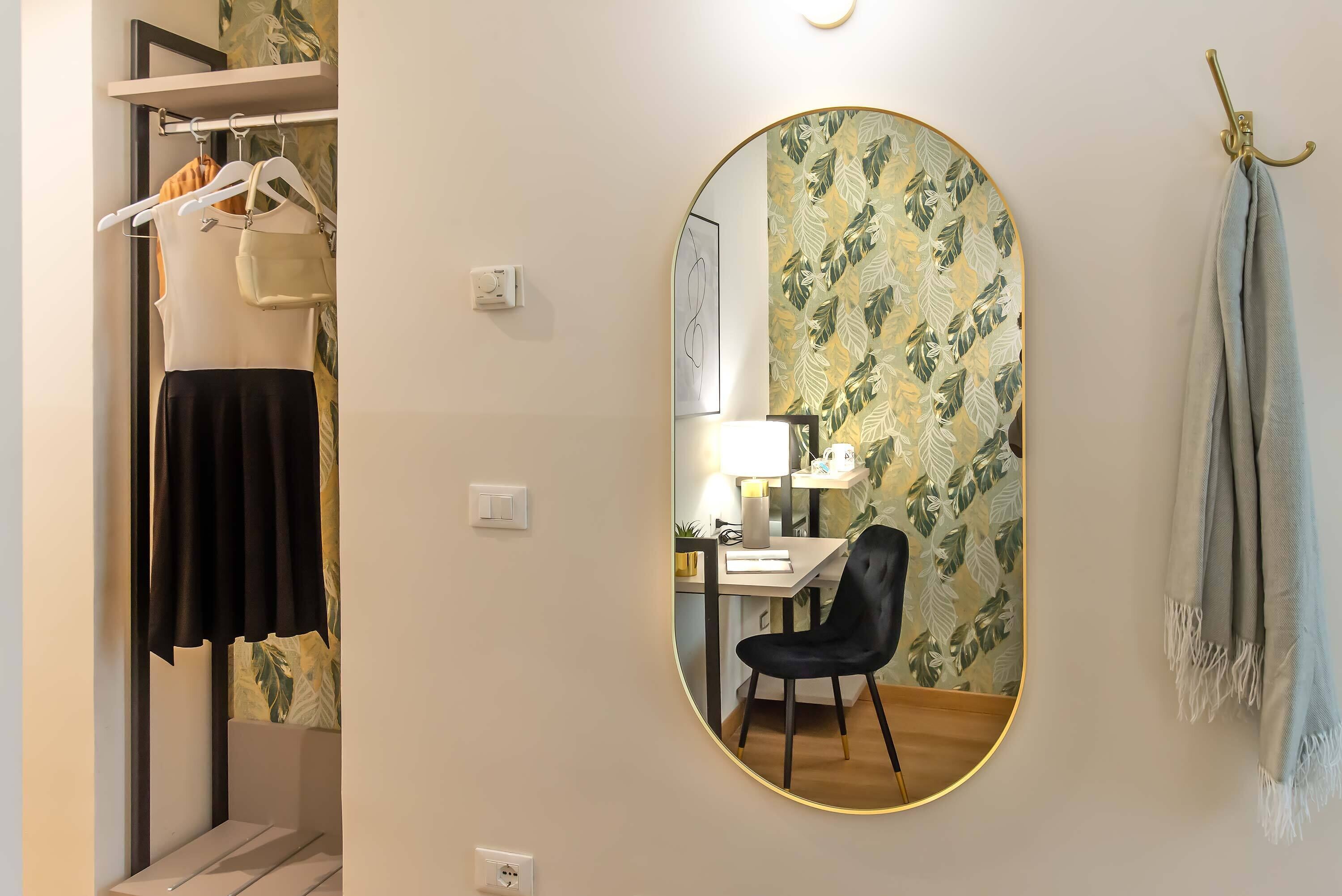 A room with a mirror, a desk with a chair, a lamp, and a wardrobe with hanging clothes.