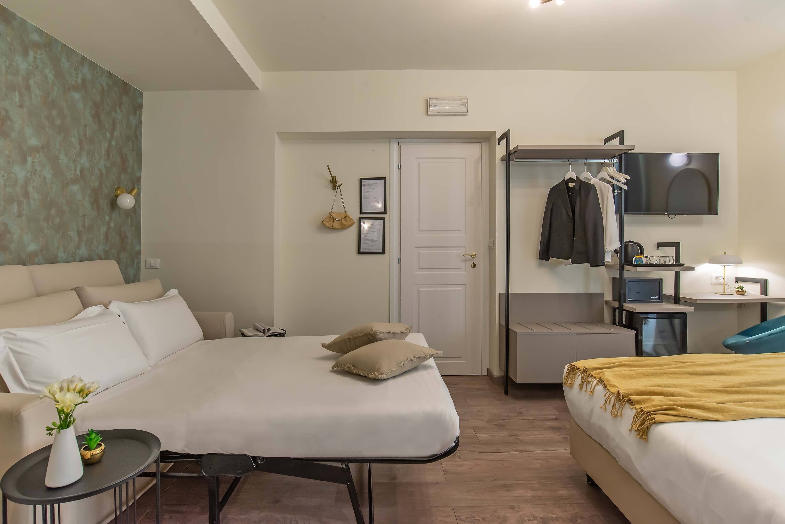 A modern hotel room with a large bed, a desk with a computer, a TV mounted on the wall, and a wardrobe with hanging clothes.