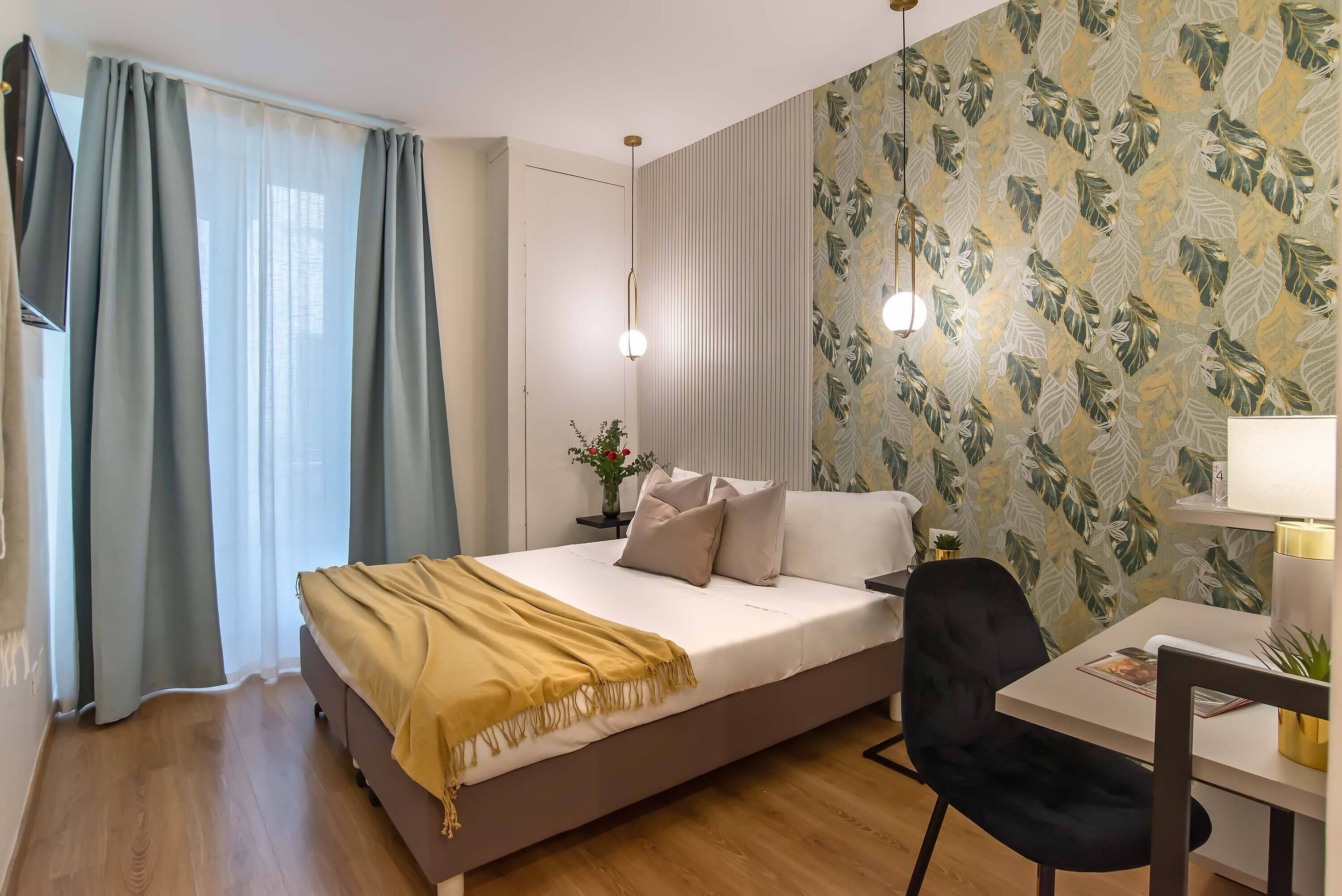 Una camera da letto con un letto, una scrivania e una sedia. Si vede una finestra con tende, una parete con carta da parati a motivi fogliacei e una pianta sulla scrivania.