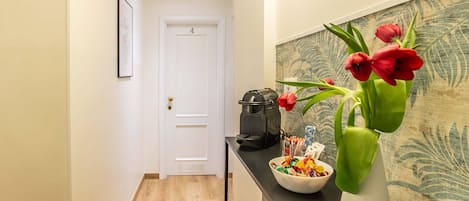 Un corridoio con pavimento in legno, una porta bianca, un quadro appeso al muro, un tavolino nero con una ciotola di snack, un vaso con fiori rossi e una pianta in vaso bianco.