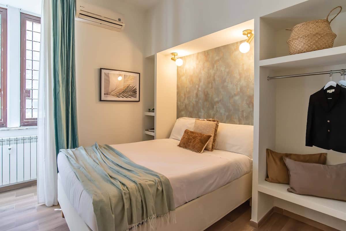 Een slaapkamer met een bed, nachtkastje, een wandplank met een gevlochten mand, kapstok en een lijst met een afbeelding aan de muur.