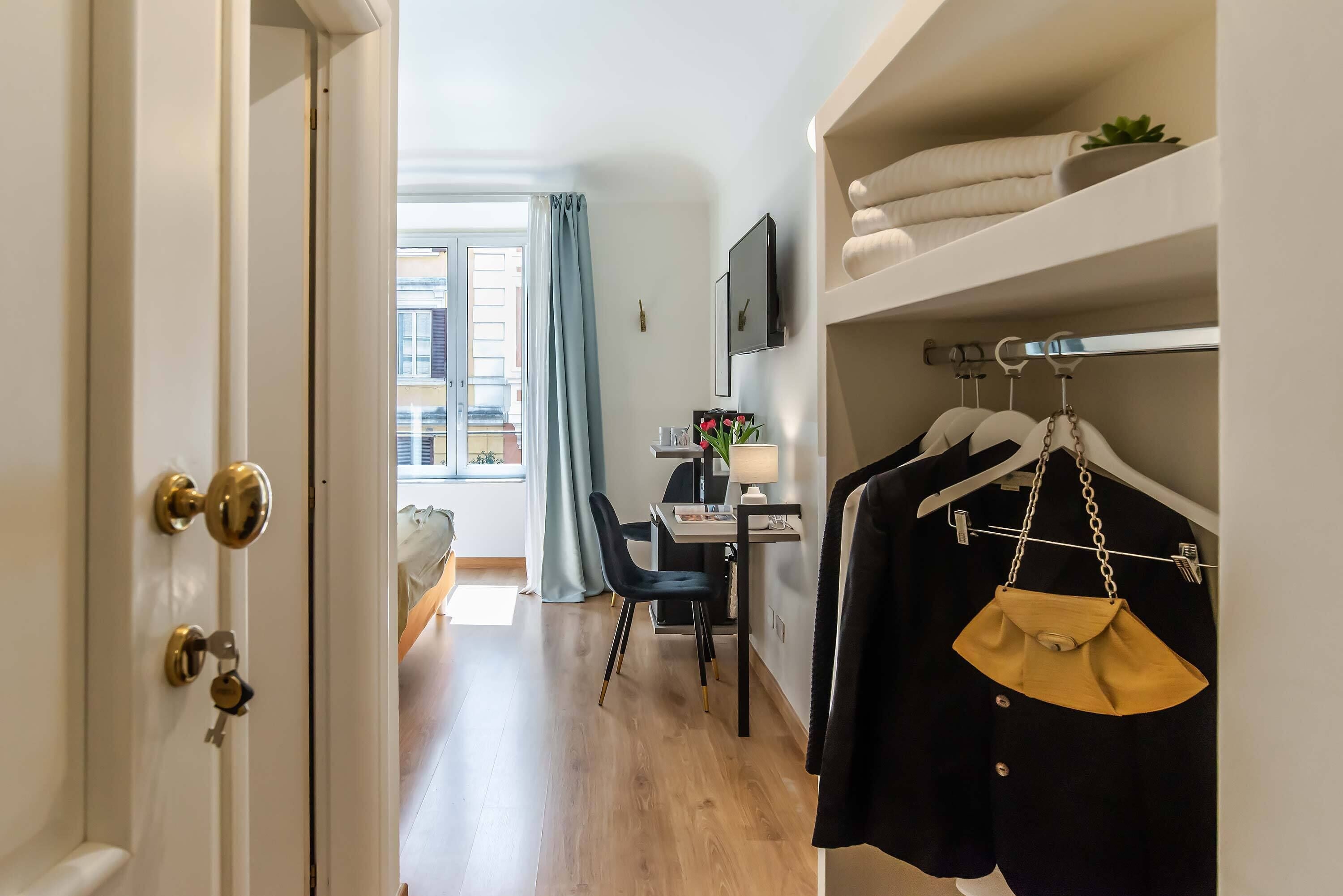Une chambre d’hôtel avec un plancher en bois, un bureau avec une chaise, un téléviseur fixé au mur et un porte-manteau avec un sac à main.