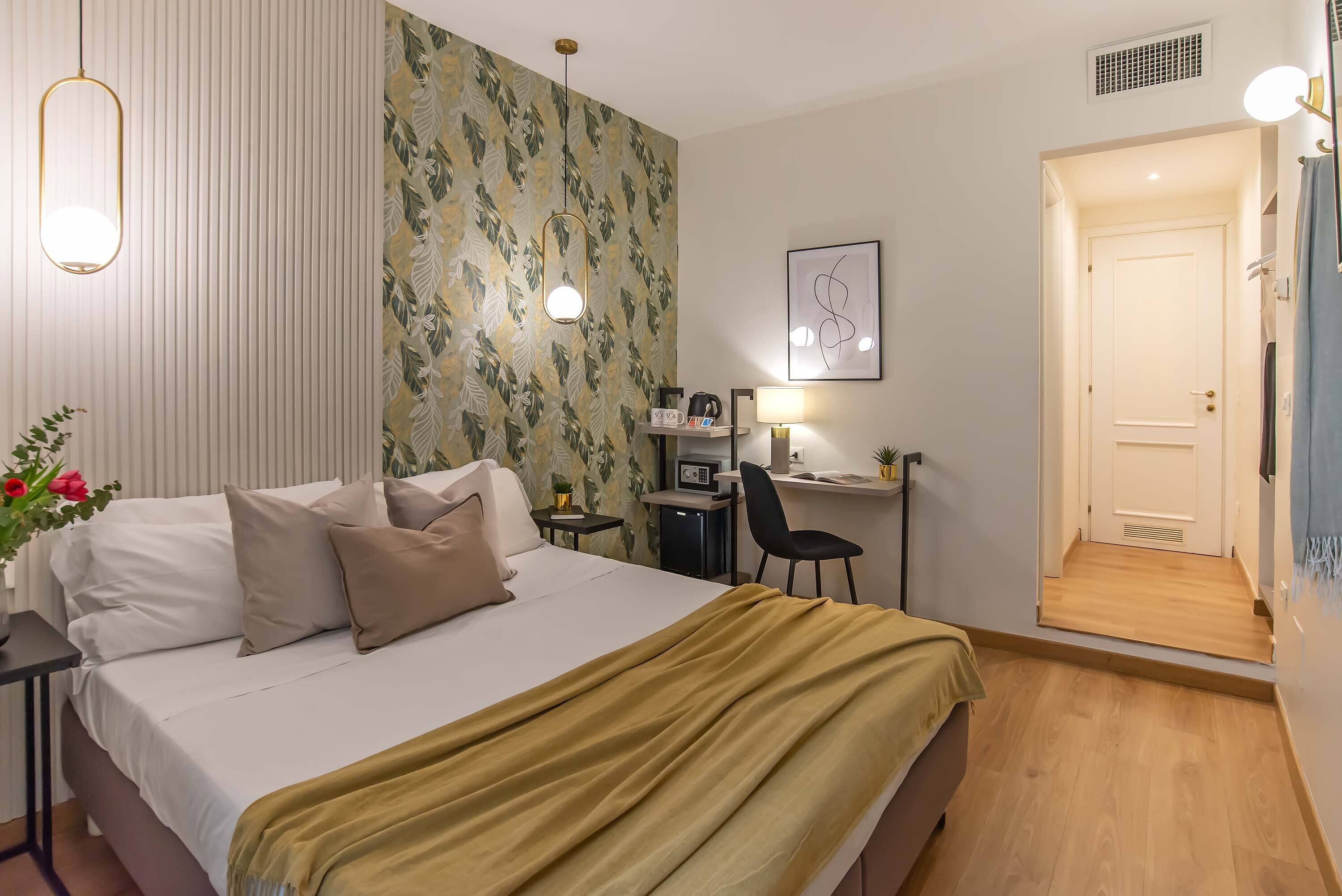 A hotel room with a bed, bedside table, desk, chair, and a door leading to another room.