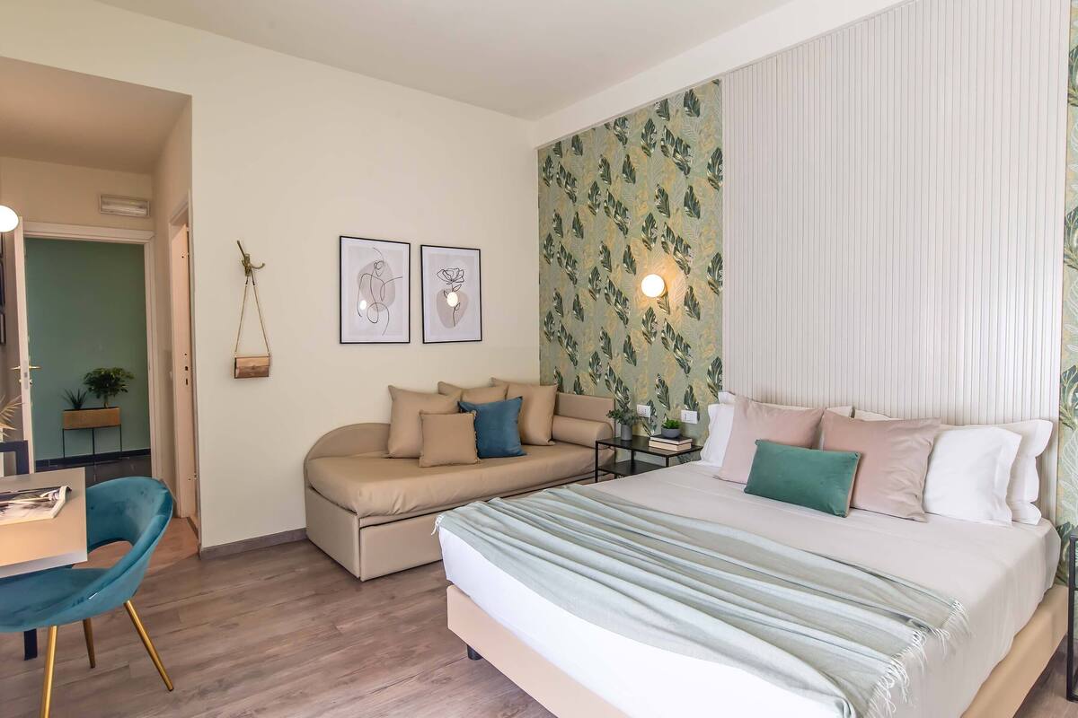 Een moderne slaapkamer met een groot bed, een bank, een bureau en een stoel. Twee kunstwerken met lijst hangen aan de muur en er staat een plant op de bank.