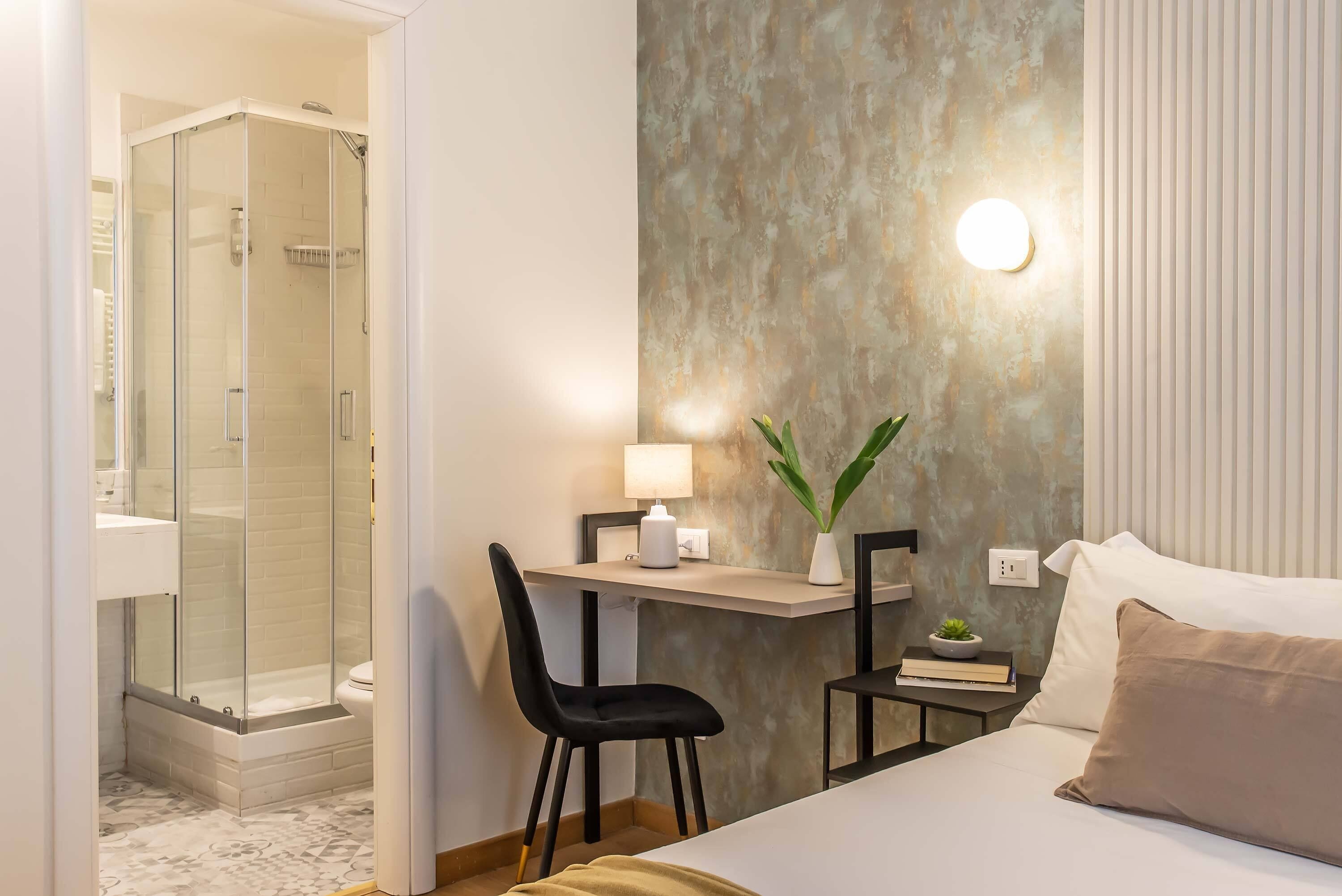 A hotel room with a shower, a desk with a chair, a bedside table with a lamp, a plant, and a bed with pillows.