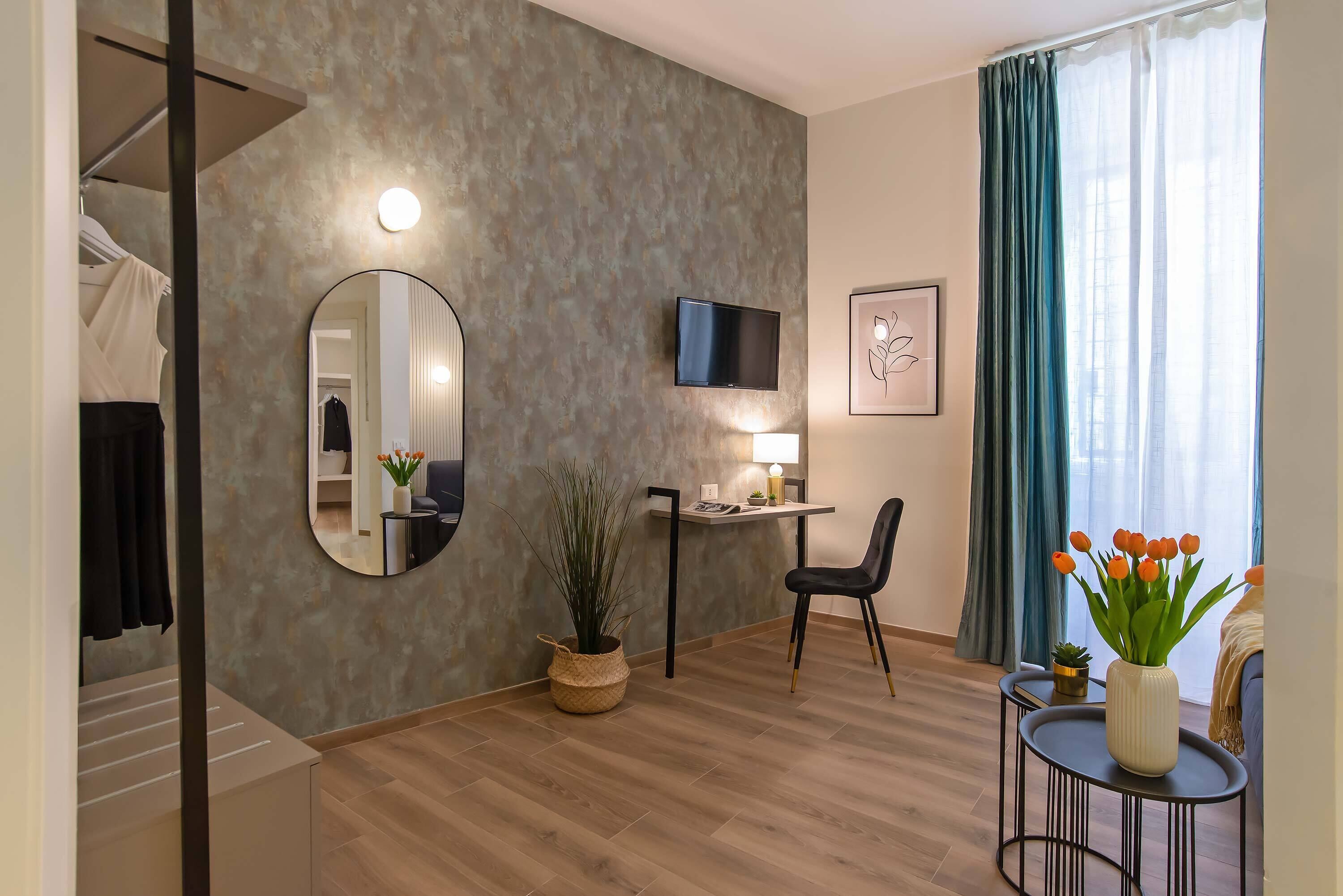 Una habitación de hotel moderna con piso de madera, un espejo redondo, un televisor de pantalla plana, un escritorio con lámpara, una silla, una mesita con un jarrón de flores y un ventanal con cortinas.
