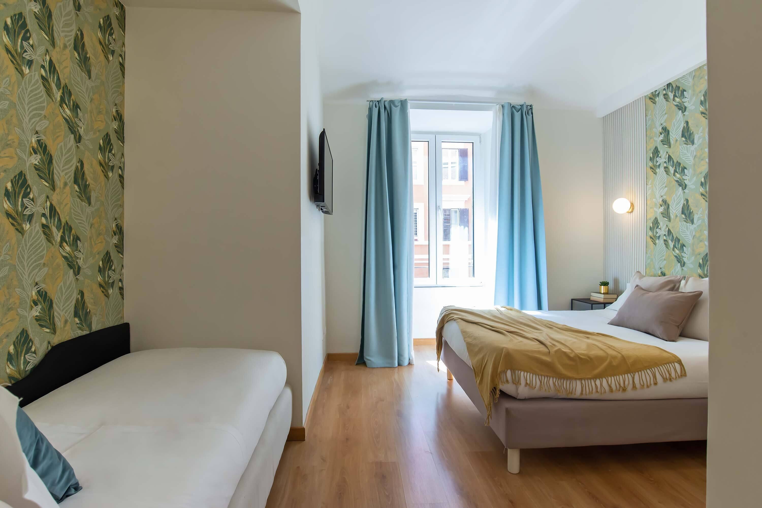 A hotel room with two beds, a TV, a window with curtains, and a small bedside table with a lamp.