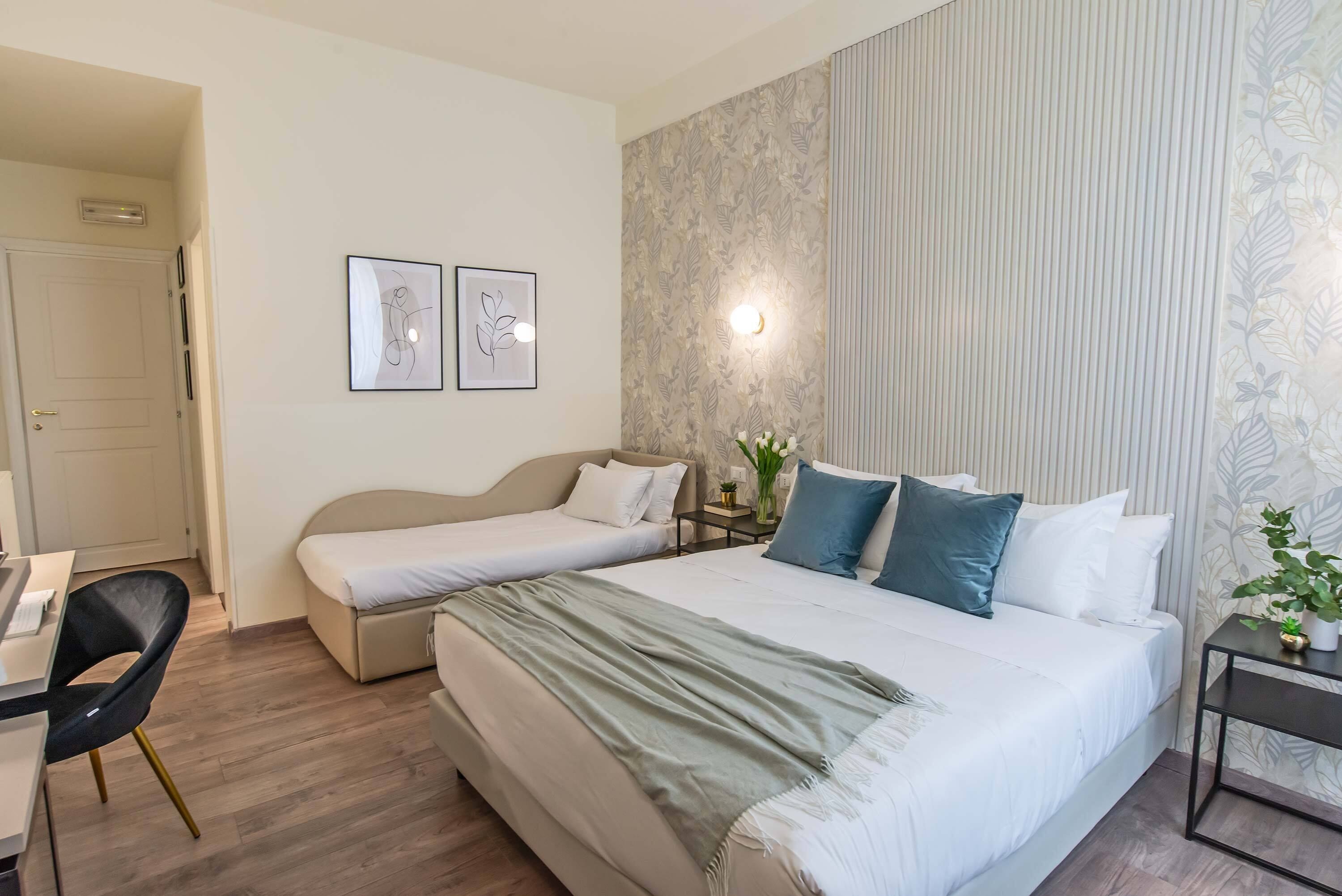 Una camera d'albergo con un letto grande, un letto più piccolo, una scrivania con una sedia e due quadri appesi al muro.