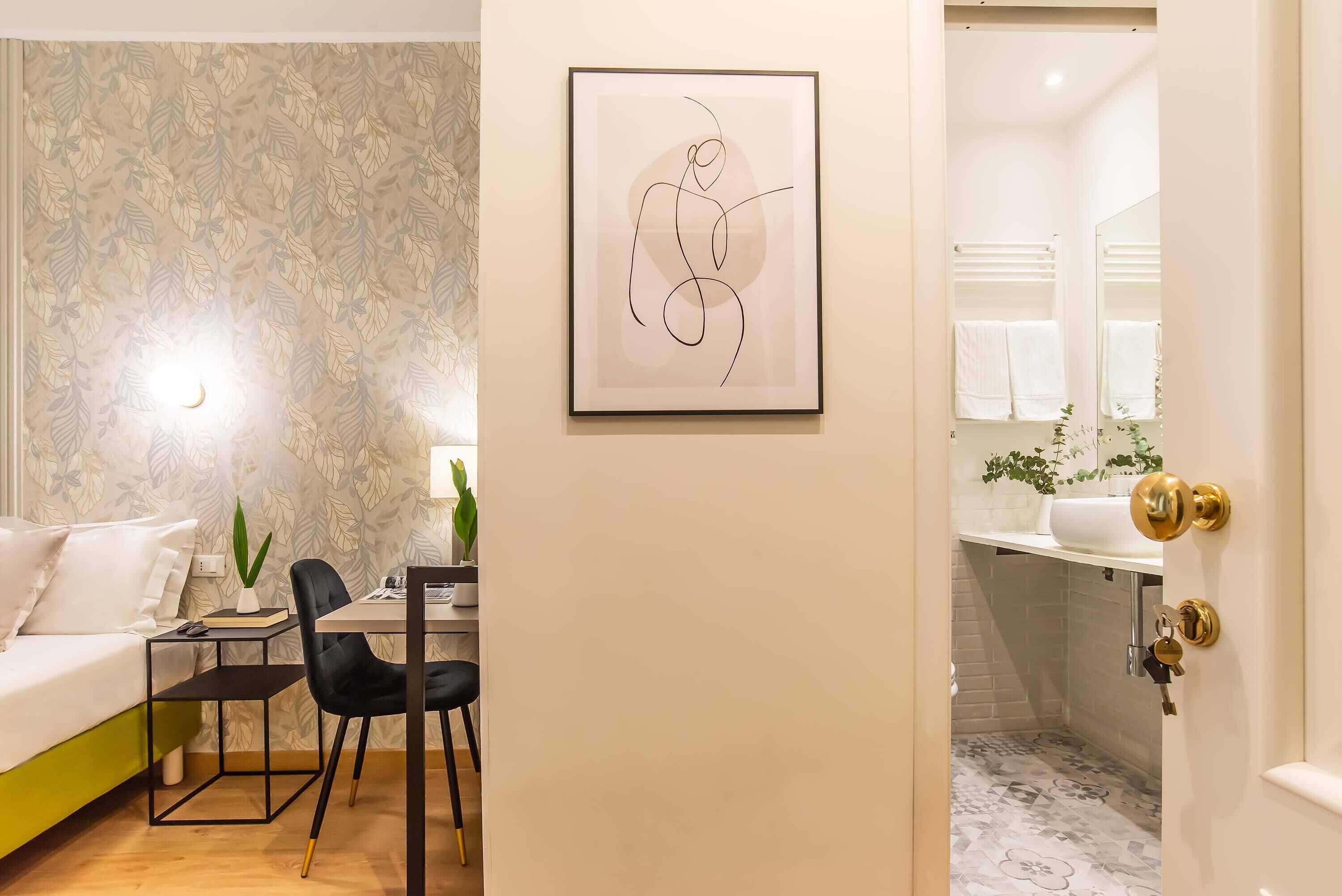 Une chambre d’hôtel moderne comprenant un lit, un bureau, une chaise, une œuvre d’art abstraite encadrée et une salle de bain avec un lavabo et un miroir.