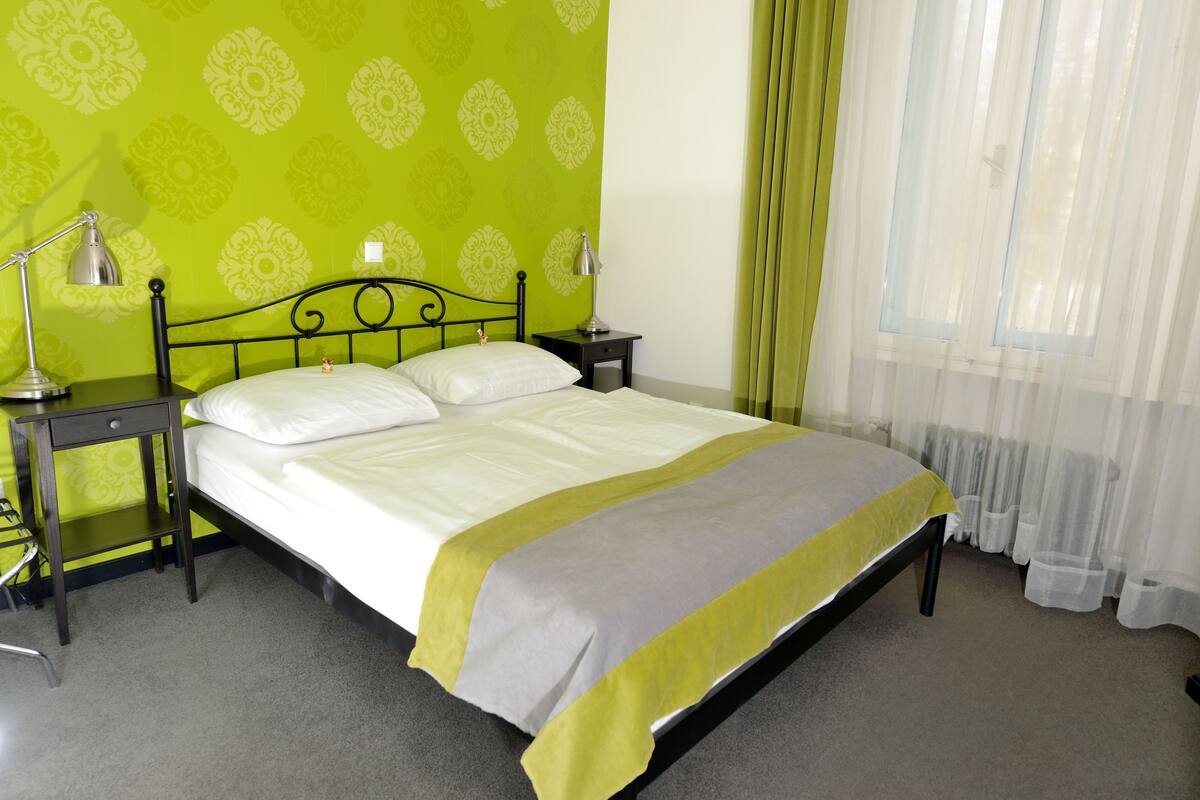 A neatly made bed with a white and grey bedspread, two bedside lamps, and a nightstand with a lamp. The room has a green patterned wallpaper and a window with sheer curtains.