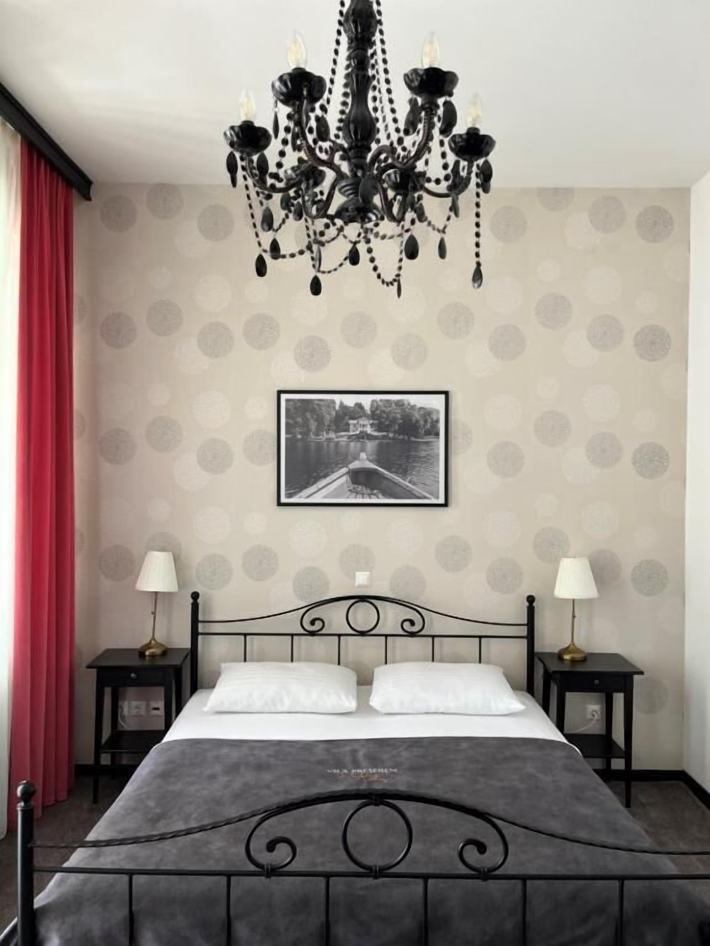 A hotel room with a bed, two bedside tables, a chandelier, a wall-mounted picture, and a red curtain.