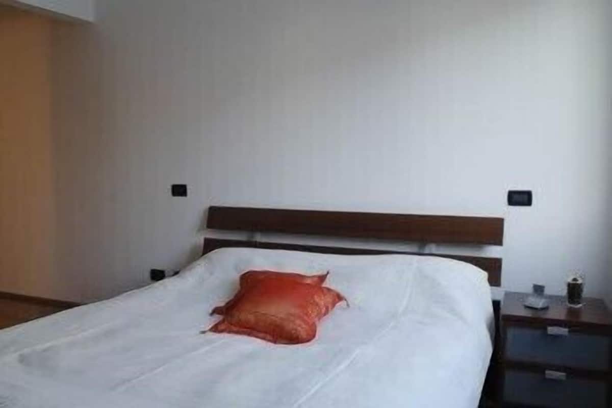 Una cama individual con cabecera de madera, ropa de cama blanca y una almohada roja. Hay una mesita de noche con una lámpara y un buró con una lámpara.