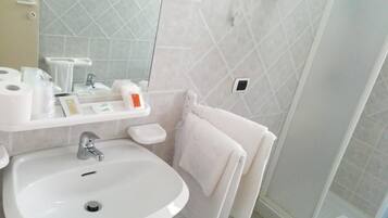 Ein Badezimmer mit weißem Waschbecken, Toilette und Dusche. Auf dem Waschtisch stehen Toilettenartikel, es gibt einen Handtuchhalter und einen Spiegel.