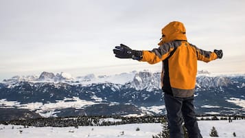 Uma pessoa com jaqueta amarela em uma montanha nevada, com os braços abertos, admirando uma cadeia de montanhas cobertas de neve.