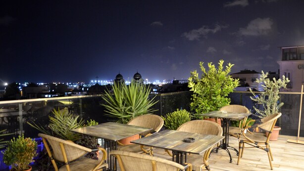 Un área de estar al aire libre con mesas y sillas, rodeada de plantas en macetas, con vista a un paisaje urbano nocturno.