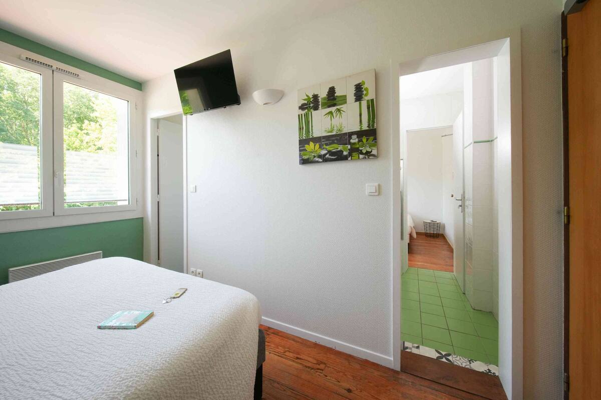 A bedroom with a bed, a TV mounted on the wall, a painting, and a view of a green-tiled hallway.