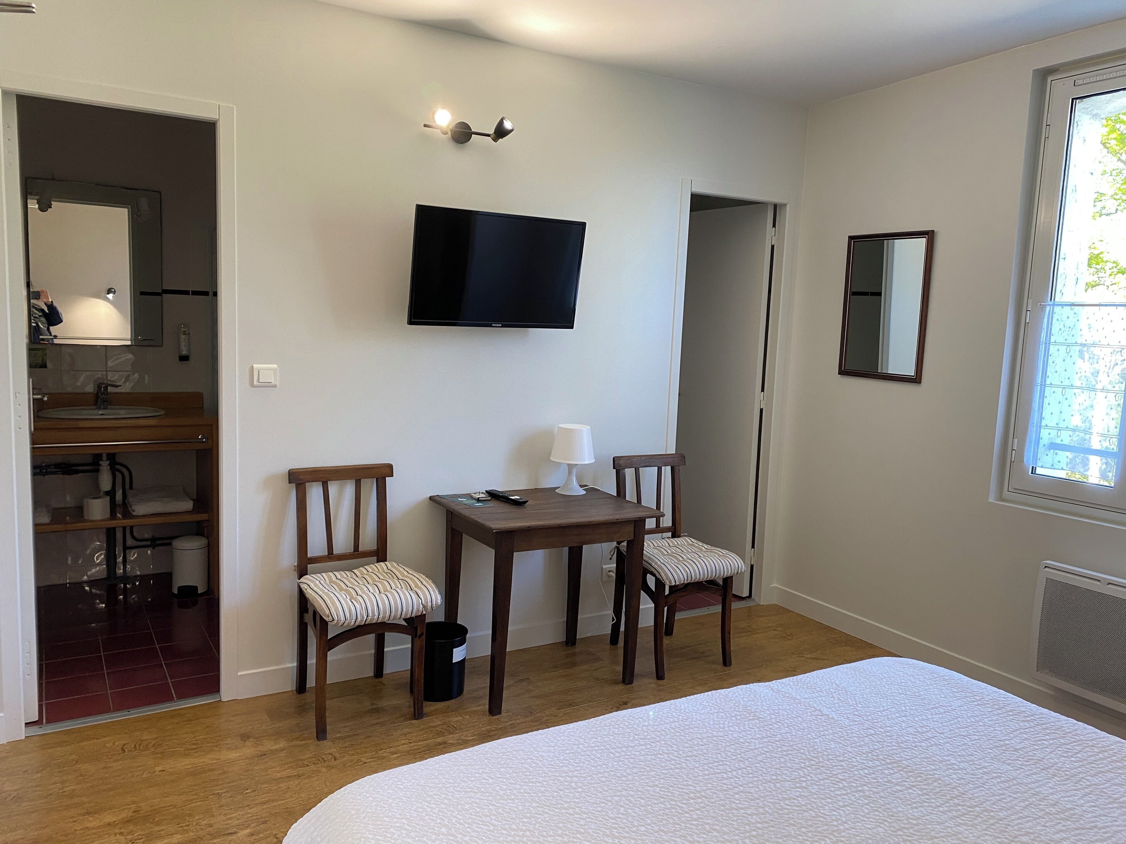 A bedroom with a bed, a TV mounted on the wall, a small table with chairs, a mirror, and a window.