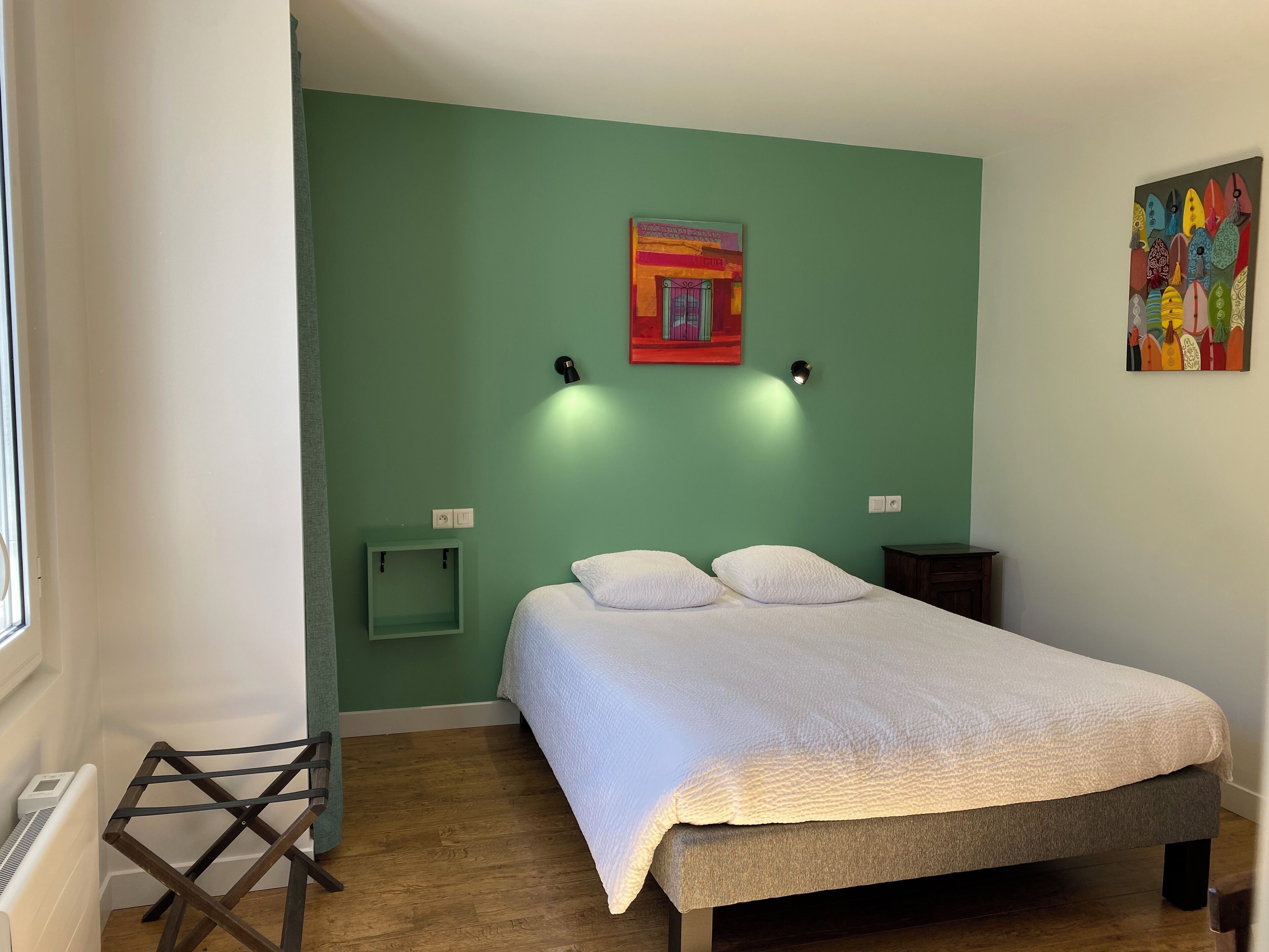 Un dormitori amb un llit, dues làmpades penjades a la paret, una petita prestatgeria verda i un quadre de colors a la paret.