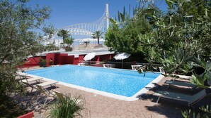 Una piscina con sillones de descanso y sombrillas, rodeada de vegetación y un edificio rojo con una estructura blanca al fondo.