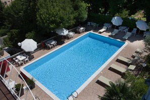A rectangular swimming pool surrounded by lounge chairs and umbrellas, with a tiled deck and greenery in the background. - Du Parc Hotel (Gabicce Mare)