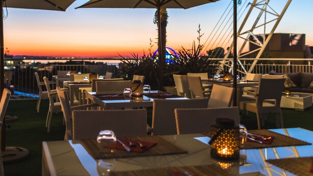 Un espacio de comedor al aire libre con mesas y sillas, sombrillas y vistas al atardecer sobre el agua.