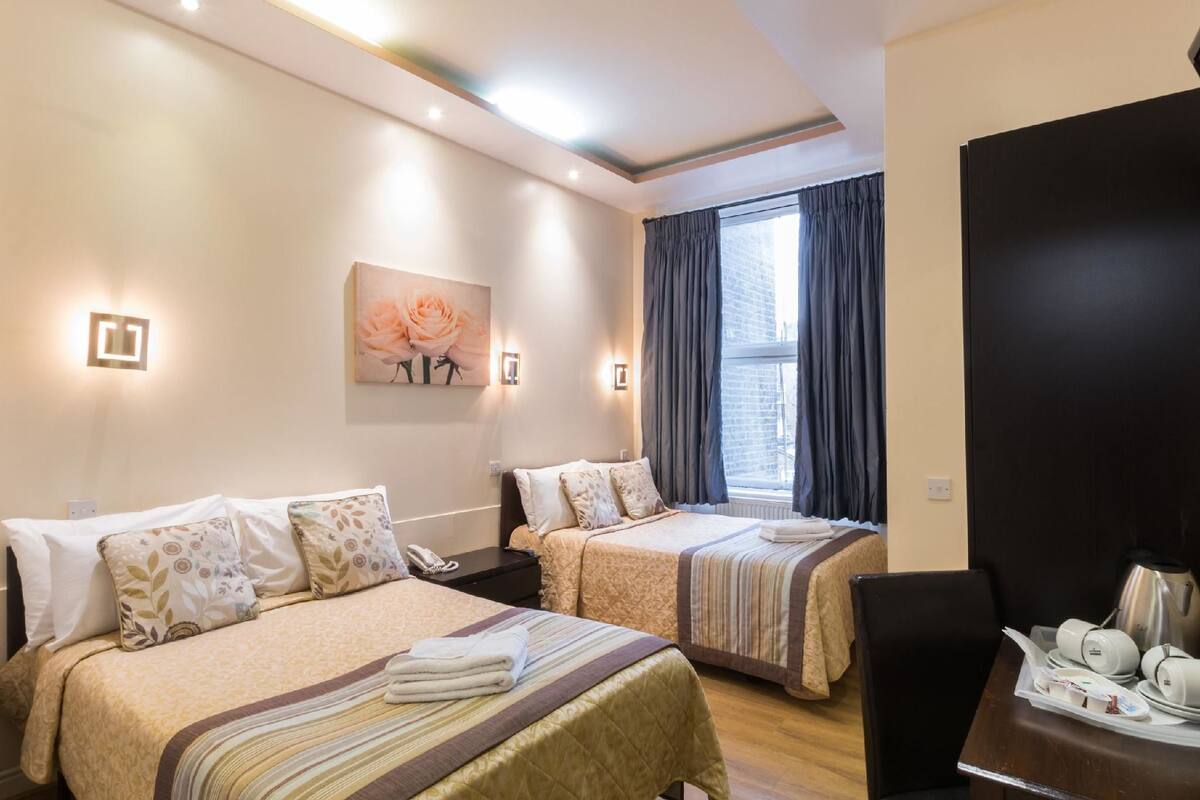 A hotel room with two beds, a painting of roses, a window with curtains, a coffee maker, and a tray with cups and a plate.