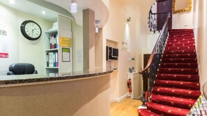 A reception area with a clock, a counter, a staircase with red carpet, and a fire extinguisher. - Notting Hill Gate Hotel (London)