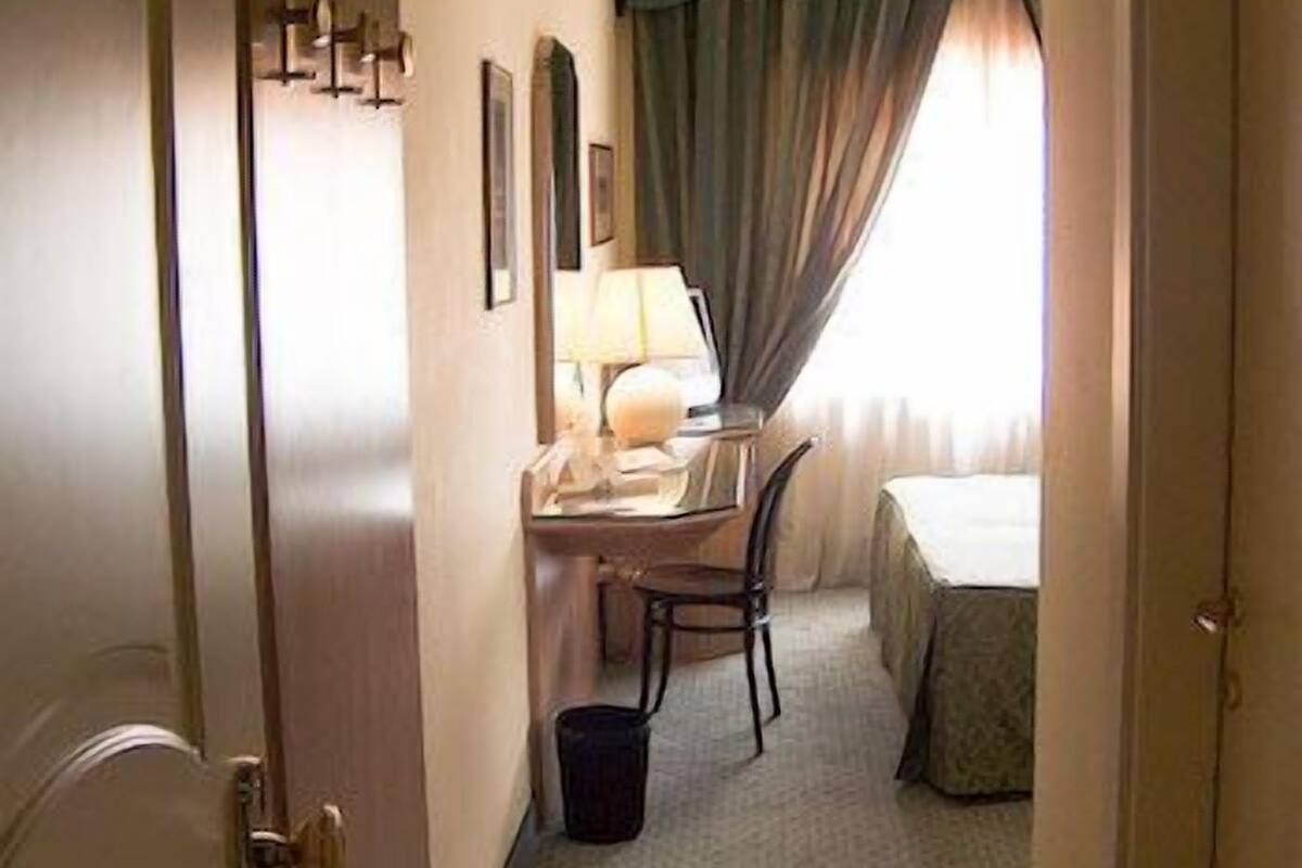 A hotel room with a bed, a desk with a lamp, a chair, and a small trash bin.