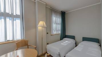 Ein Hotelzimmer mit zwei Einzelbetten, einem Tisch, einem Stuhl, einer Stehlampe und einem Fenster mit Vorhängen.