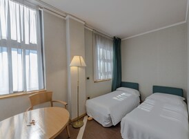 Ein Hotelzimmer mit zwei Einzelbetten, einem Tisch, einem Stuhl, einer Stehlampe und einem Fenster mit Vorhängen.