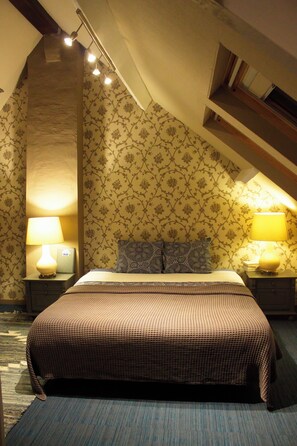 Un dormitorio con cama, dos mesitas de noche, una lámpara en cada una, pared con diseño y techo inclinado.