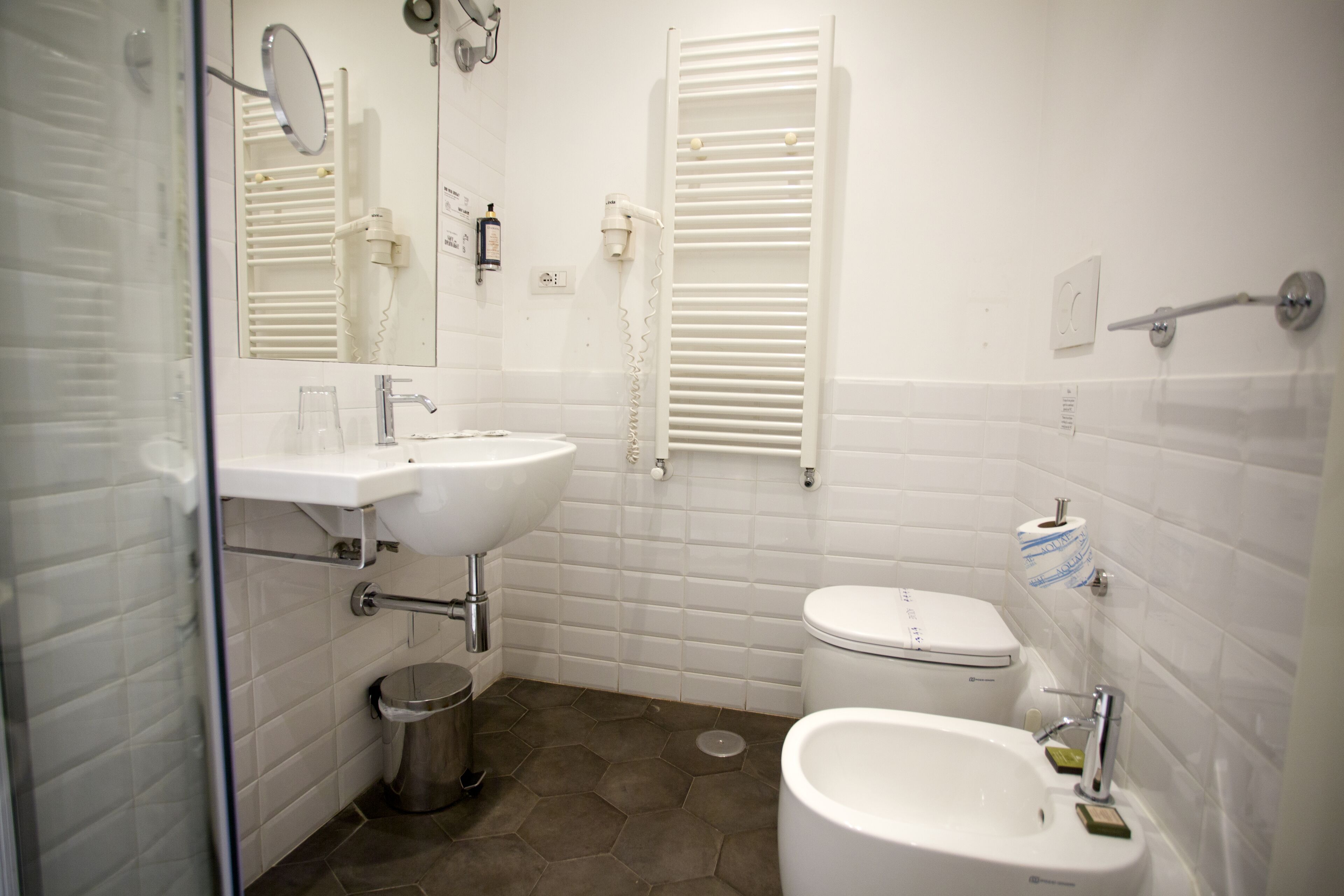 A modern bathroom with a white sink, toilet, and bidet, a glass shower enclosure, and a trash can.