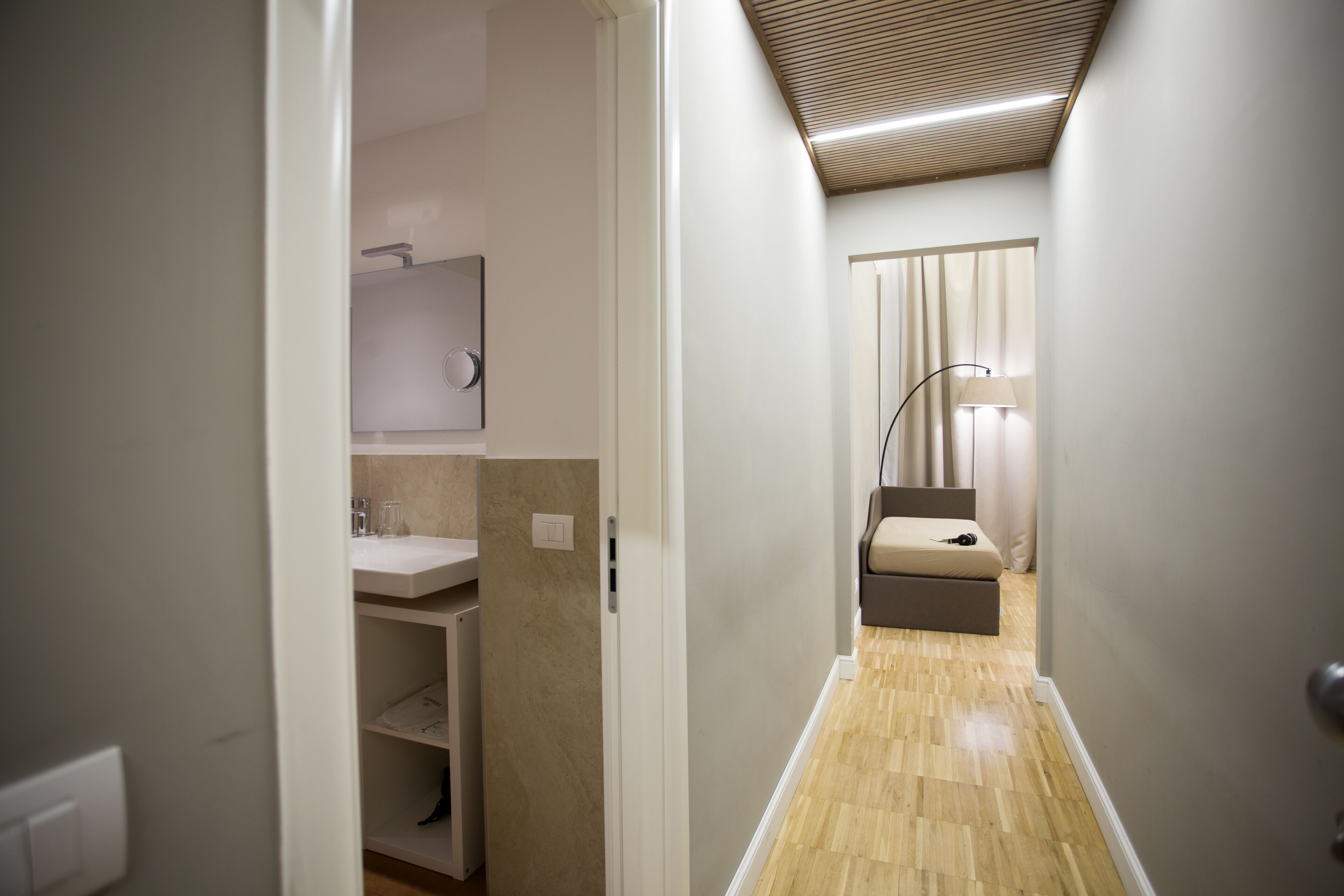 A narrow hallway with a wooden floor, a white door on the left, and a bed with a headboard and curtains on the right.