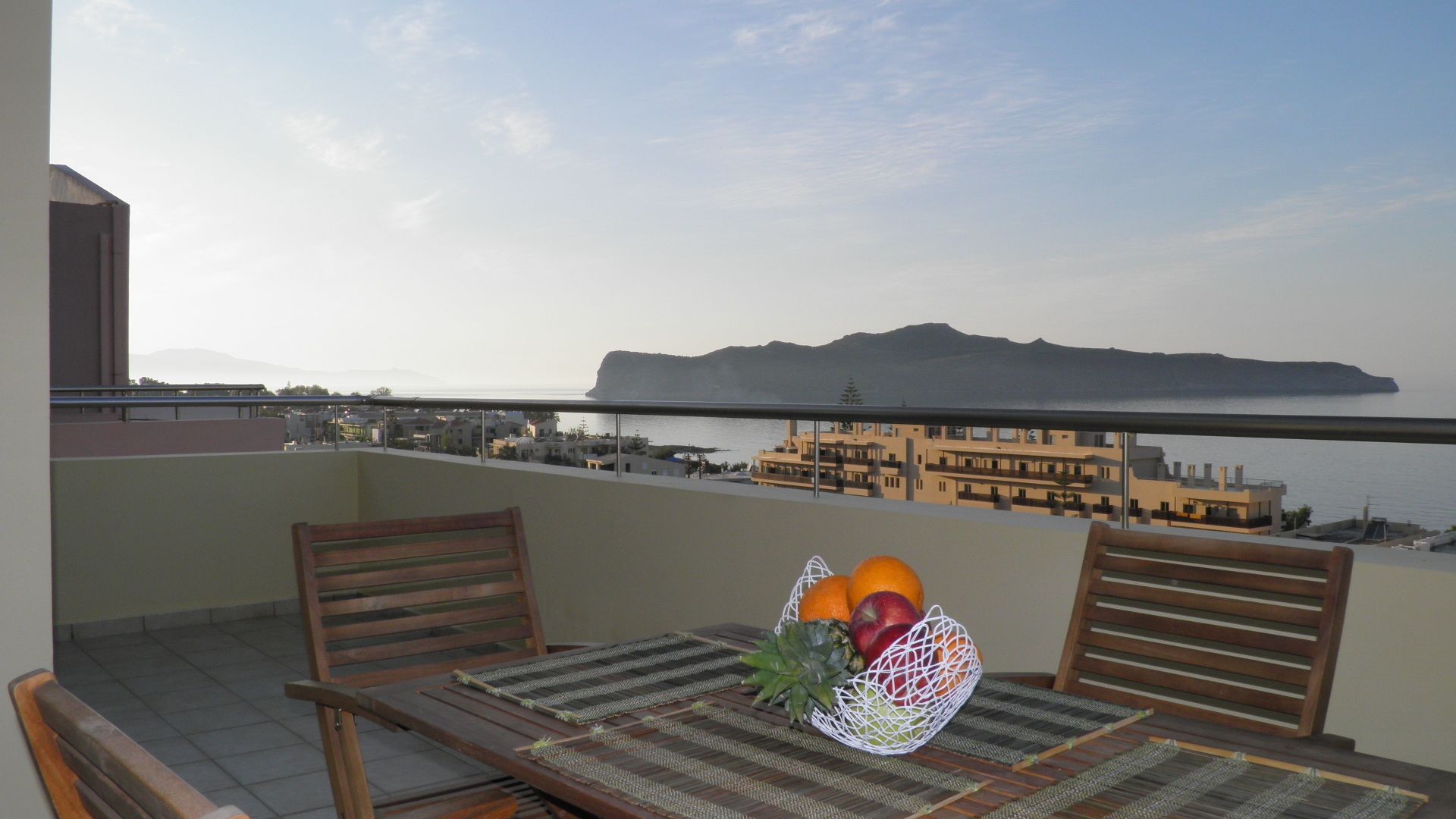 A balcony with a table set for two, featuring a woven placemat and a fruit basket with oranges and apples.