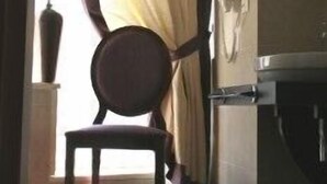 Ein Zimmer mit einem Sessel in der Nähe eines Fensters mit Vorhängen, einer Topfpflanze und einer Wandleuchte.